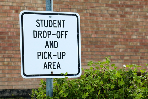 Image of a sign that says this is the student drop off and pick up area at a school.  Behind the metal sign is a brick wall and beside it is a bush.