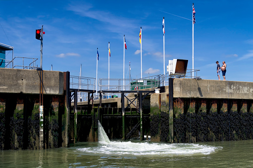 Shotley Gate, Suffolk, England - May 27, 2015: Water pouring through the lock gates at Shotley Gate marina as they prepare to open to allow a boat through into the harbour. A man and a youth are watching as they fish from the quayside with handlines.