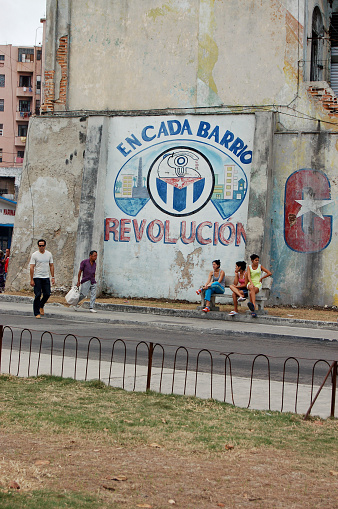 Havana, Cuba - November 18, 2005:  Local residents sitting and walking by a mural marking the Communist Revolution on a cloudy afternoon  in Old Havana, Cuba