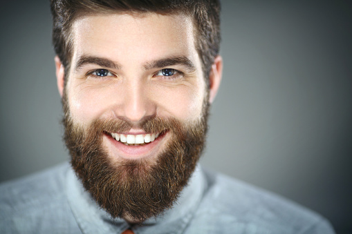 Closeup portrait of smiling handsome early 30's man with blue eyes.He has neat brown hair and fully grown beard.Wearing gray shirt.Looking at camera with a toothy smile.