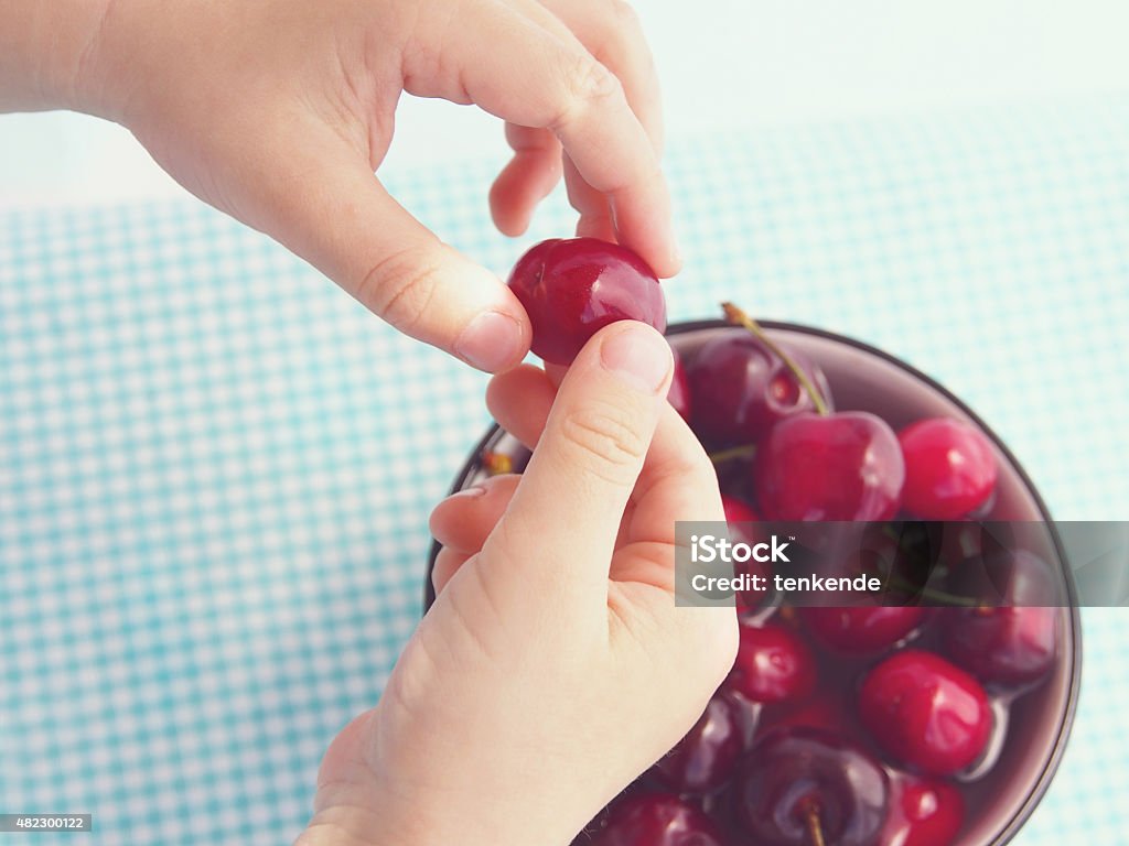 Child eating cherries Cherries in a ceramic bowl on checkered table cloth with  hands of a child keeping one of the cherries above the bowl 2015 Stock Photo