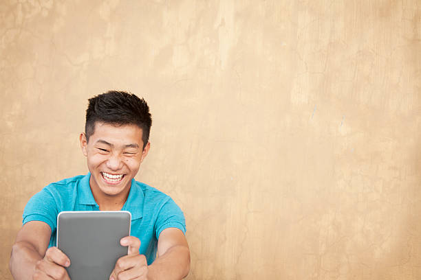Education. Asian teenage boy laughs while using digital tablet. Back to school. Asian, Indian descent teenage boy enjoys using his digital tablet to study. He holds the tablet and laughs while he learns more about his world via the internet. Close-up portrait style image. High school or college age student. secondary school activities stock pictures, royalty-free photos & images