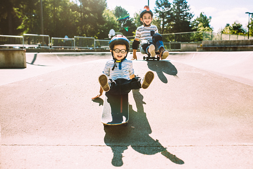 Two fun, playful Hispanic boys together at a skate park. The brothers have helmets and padding, and are having fun riding down the ramps sitting on their boards. Horizontal with copy space.