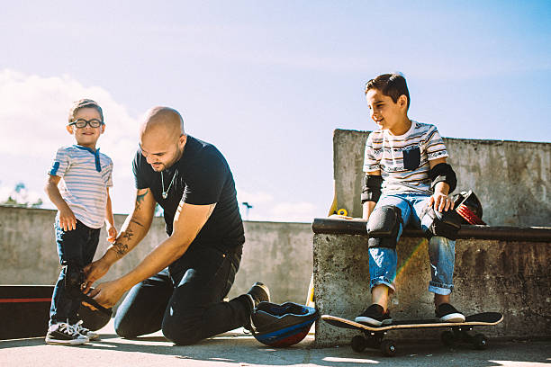 Father and Sons at Skate Park A fun, playful Hispanic Dad and his two boys play together at a skate park. The father helps his sons put on helmets and padding so they can skateboard safely.   Horizontal with copy space. kneepad stock pictures, royalty-free photos & images