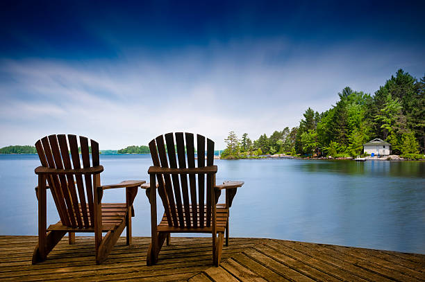 Wood Muskoka chairs on a lake deck A couple of wooden Muskoka chairs sitting on the dock with a lake and cottages across in the background. Perfect for cottage related applications cottage photos stock pictures, royalty-free photos & images