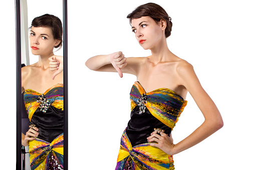 Photo of a woman wearing a dress with her thumbs down while looking at a mirror.  She is displeased with the choice of clothing and dislike the attire.  She is either a model or a snobby shopper.  The woman is isolated on a white background.