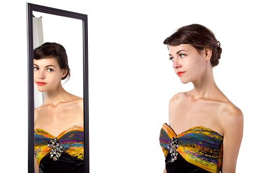 Photo of a woman wearing a yellow dress looking at her self in a mirror.  She is deciding on what outfit or attire to wear.  She is isolated on a white background.  She is either a fashion model or a shopper browsing for clothes.  She is looking at the mirror with self confidence.