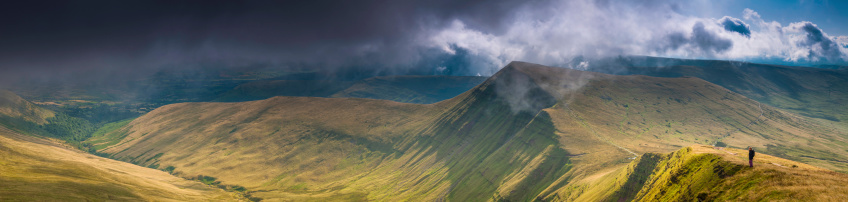 Young hiker looking out over the steep slopes and rocky ridges of Cribyn and Bryn Teg above the grassy valley of Cwm Sere under dramatic stormy skies deep in the Brecon Beacons National Park of Wales, UK. ProPhoto RGB profile for maximum color fidelity and gamut.