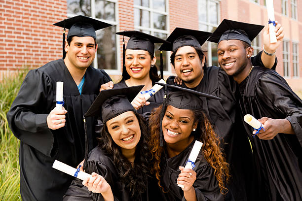 Education: Multi-ethnic friends excitedly hold diplomas after college graduation. Diverse group of friends dressed in cap and gowns excitedly show off diplomas after college graduation. School building background. mortarboard photos stock pictures, royalty-free photos & images