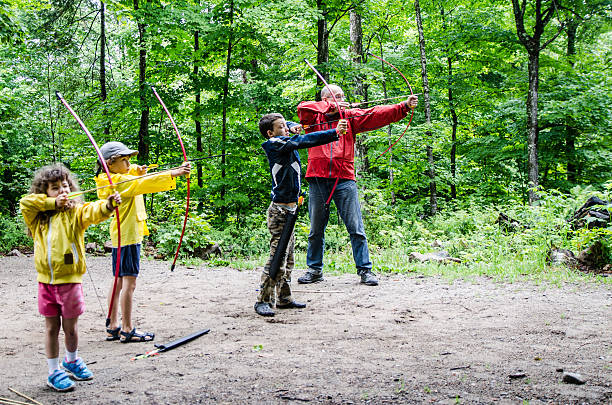 Father and children, seen from the side, using a bow to throw arrows at a target outside in the woods.