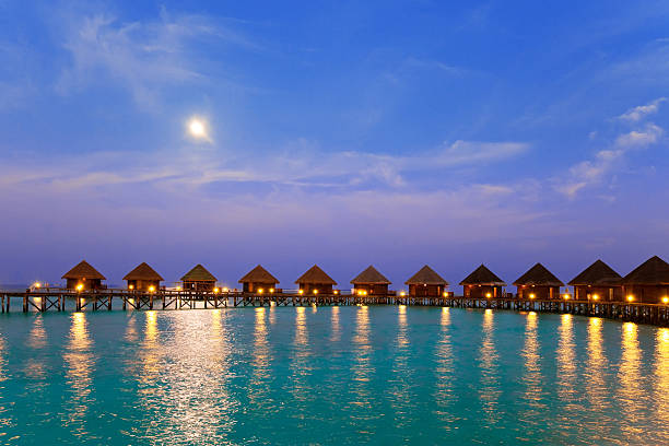 Tranquil scene of a wooden pier illuminated by lanterns, leading to a luxurious resort on a tropical island in the Maldives at dusk