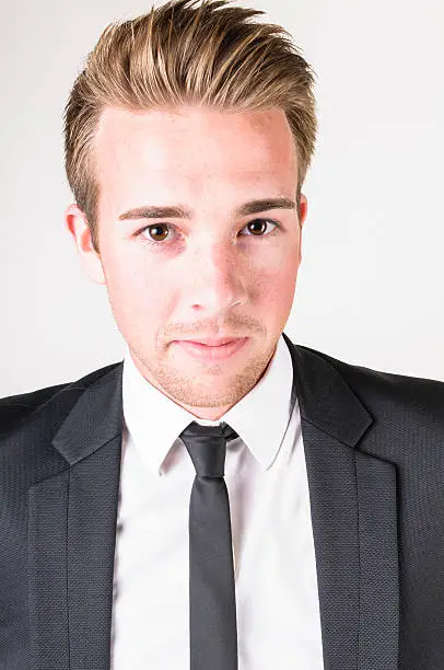Young man with blonde hair wearing a dark suit with a white shirt and is standing casually smoking a cigar in front of a white background.