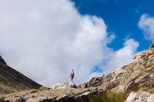 A little girl stands on a mountain ridge, all alone in the wilderness, against a lightly clouded sky.