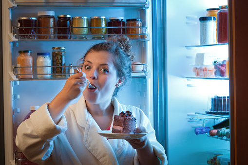 A young caucasian woman standing in front of the open refrigerator, eating unhealthy chocolate cake at late night in a domestic home kitchen. She is dressed in a casual pink shirt, looking at the camera with a surprised expression. A symbol of unhealthy eating habit and lifestyle. Photographed in vertical format.
