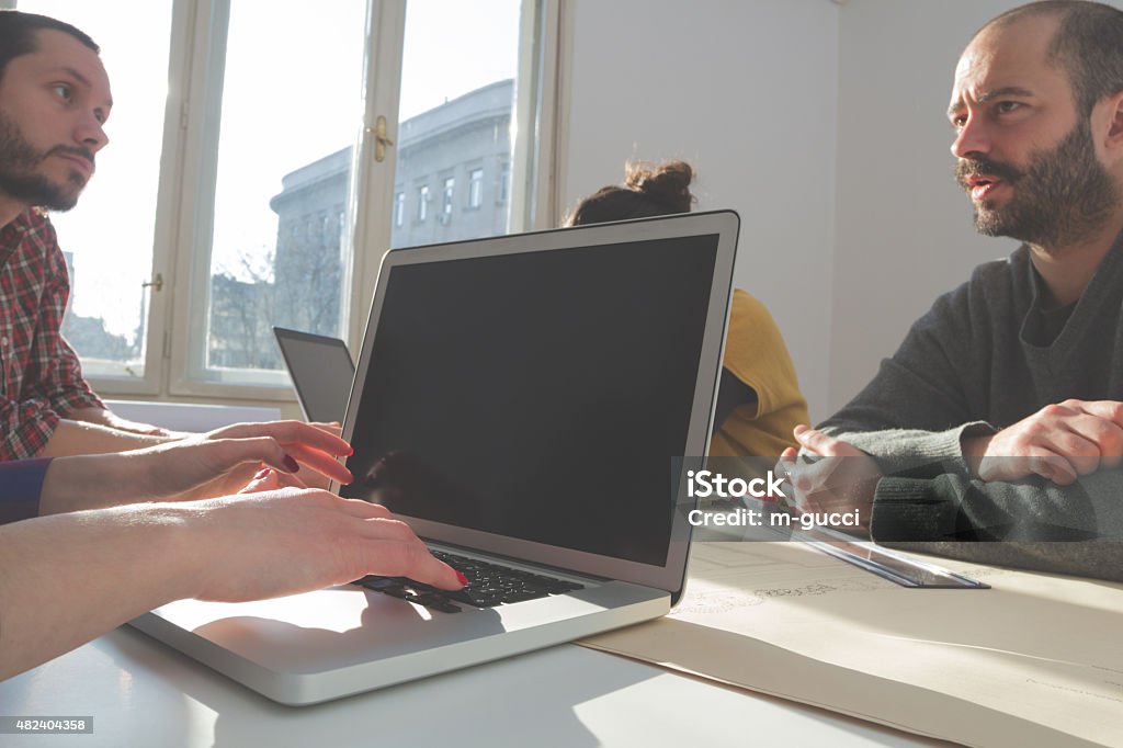 Young group of people/architects discussing business plans Young group of people discussing business plans, with lap-tops, plans in a room lit by natural sunlight. 2015 Stock Photo