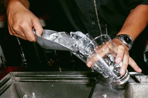 A DSLR close up of bartender putting ice cube to a glass.The bartender preparing cocktail. He is holding on 2 glasses.The bartender working behind the bar, at night in the nightclub.