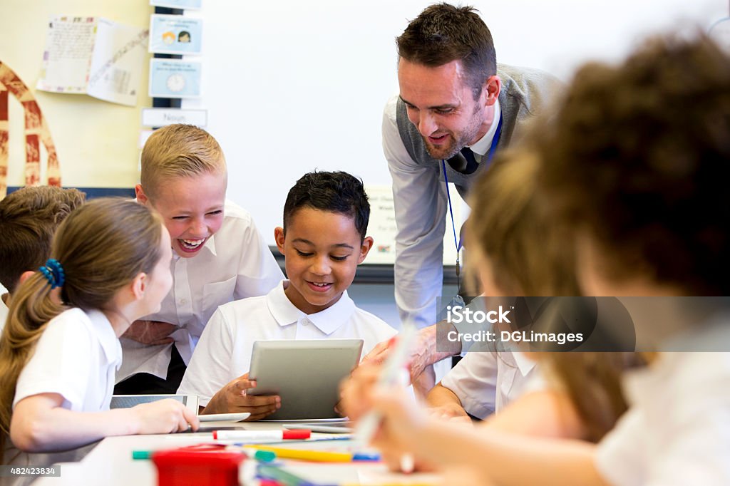 Teacher Happy at School A male teacher sits supervising a group of children who are working on whiteboards and digital tablets. Teacher Stock Photo