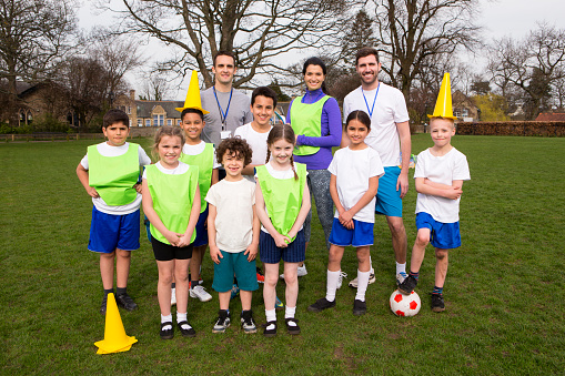 A group portrait of a kids soccer team, behind them are their coaches. They are smiling and two of the boys can be seen holding cones on their head messing amount.