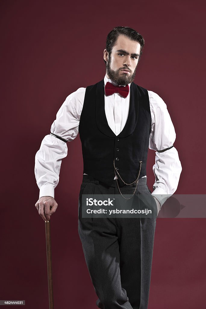 Retro 1900 victorian fashion man with beard wearing black gilet. Retro 1900 victorian fashion man with beard wearing black gilet and red bow tie. Holding a walking stick. Studio shot against red wall. Adult Stock Photo