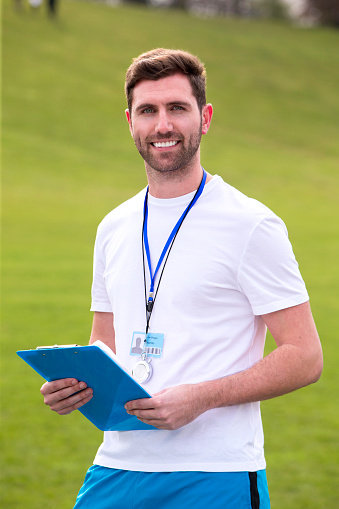 A male sports teacher stands outdoors on a field, he is holding a clipboard.