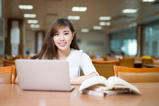 Beautiful asian female student using laptop for study in library with bookshelf background.