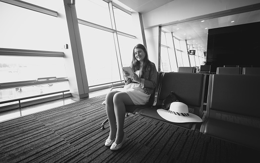 Black and white photo of young woman sitting in airport terminal and using tablet