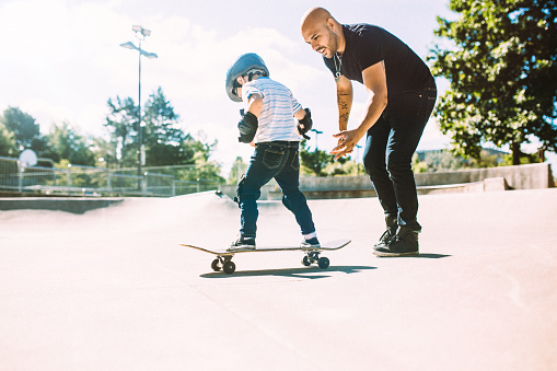 A fun, playful Hispanic Dad and his son play together at a skate park. They boy has a helmet and padding, and is having fun with his father. The father smiles as he teaches his young boy to skateboard, holding his arms for balance. Horizontal with copy space.