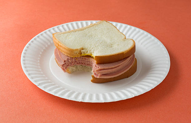 Large bitten bologna sandwich with mustard A bitten bologna sandwich with mustard and white bread on a paper plate atop an orange table top. baloney photos stock pictures, royalty-free photos & images