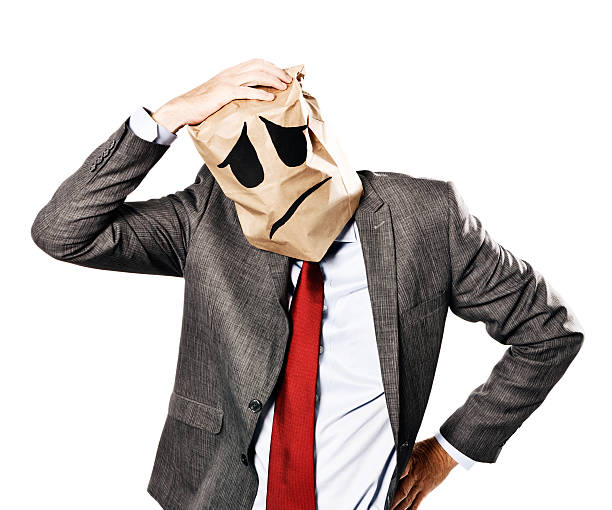 Businessman in depressed-looking paper-bag mask scratches his aching head A mature businessman, in formal business clothes, wearing a mask made from a paper bag, showing a downturned, unhappy mouth scratches his head, depressed, confused, and probably with a headache! anonymous businessman with paper bag on his head stock pictures, royalty-free photos & images