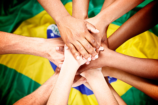 Many mixed hands are piled up in unity against a background of the Brazilian flag, in patriotic support, of the nation or perhaps a sports team.