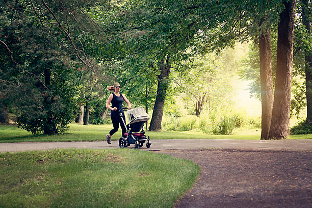 Getting back to shape after baby! This new mother is jogging while baby is sleeping in the stroller. They are in a public city park on a sunny summer day. Full length horizontal shot with copy space and sun flare. This was taken in Montreal, Canada.
