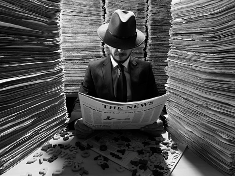 Mid adult man sitting on desk in low key lighting and reading newspaper.Large amount of newspapers are stacked on desk and on the background.He is wearing a black suit, necktie and a fedora hat.Up half of his face is obscured with Hat.The photo was shot with medium format DSLR camera Hasselblad and a wide angle lens in studio lighting.