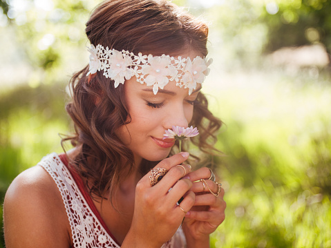 Boho teenager in a sunlit summer park serenely smelling a small pink daisy flower, her eyes closed with pleasure
