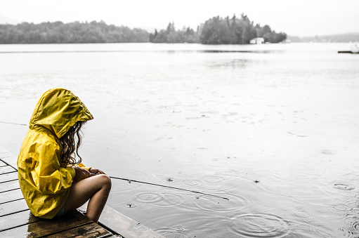 A little girl wearing a yellow raincoat is sitting on the dock and fishing in the lake Sept-Iles (Portneuf) while it's raining.