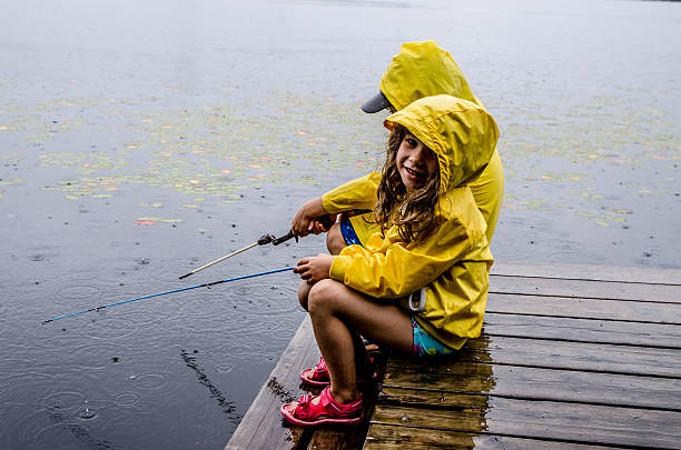 黄色いレインコートを着た小さな女の子と小さな男の子がドックに座り、雨が降っている間に湖の9月イル(ポルトヌフ)で釣りをしています。