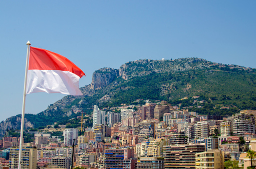 National flag of Monaco with the city Monte Carlo and town houses in background with blue sky and copy space. This travel colour image is a symbol for the microstate on France’s Mediterranean coastline known for its casinos, yacht-lined harbor and Grand Prix motor race. Monte Carlo, its major district, has an elegant casino complex, ornate opera house and luxe hotels, boutiques, nightclubs and French and Italian restaurants.