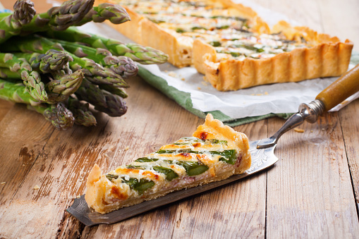 Slice of delicious home made asparagus quiche with pecorino and bacon on rustic wooden background. Selective focus.