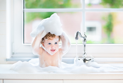 Funny baby girl playing with water and foam in a big kitchen sink next to a window