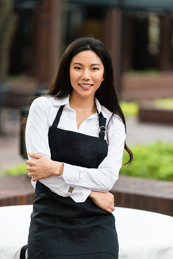 Asian waitress leaning against table at outdoor restaurant with arms crossed