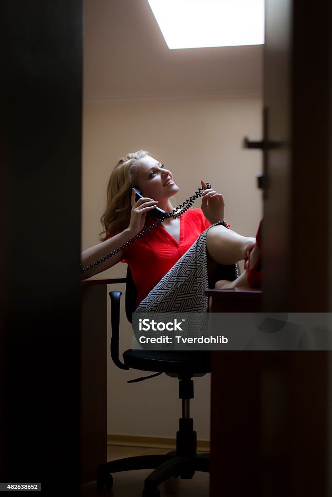 Beautiful female secretary with phone View from doorway of smiling beautiful blonde secretary woman sitting in office with feet on table in red blouse holding telephone receiver speaking on phone, vertical picture 2015 Stock Photo