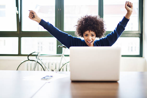 Young Woman Celebrating Success in Loft Space Behind Laptop Young black woman celebrating success in a modern loft space behind a laptop. super bike stock pictures, royalty-free photos & images