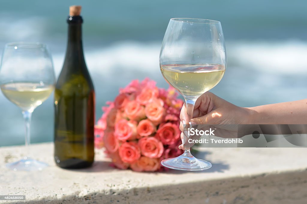 Wedding bouquet bottle and wine glasses Closeup of female hand green bottle one wedding coral rose bouquet and two glasses with white wine standing on parapet sunny day outdoor on wavy blue ocean or sea water background, horizontal picture Coral Colored Stock Photo