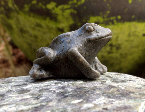 Photo showing a grey marble frog decoration, perched on top of a large pebble, with green lichen providing the backdrop.