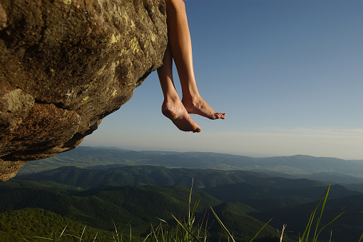 Beautiful landscape view from high mountaing on green hills with wood and human female legs barefoot of girl sitting on rock on natural blue sky copyspace background, horizontal picture