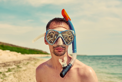 funny portrait of man on beach with swimming goggles with sand inside it, looking through camera, vacation concept.
