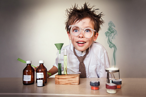 A young boy is dressed as a mad scientist.  There are bottles and beakers in front of him, and he is wearing glasses.