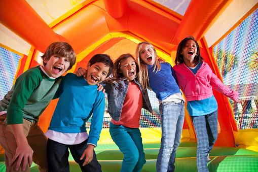 A group of five young children laughing in an orange, yellow and green bounce house.  The children are wearing colorful clothing and have their arms around each other.