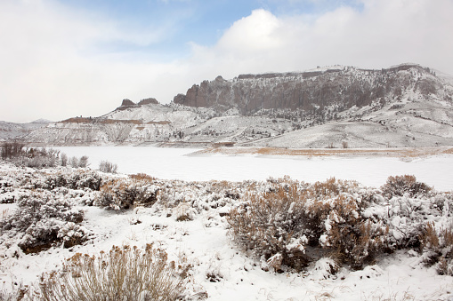 The snow covered Dillon Mesa and volcanic Dillon Pinnacles stand behind an ice and snow covered Blue Mesa Reservoir, part of the Curecanti National Recreation Area along the Gunnison River outside Montrose, Colorado. Rabbitbrush and sagebrush stand in the foreground.