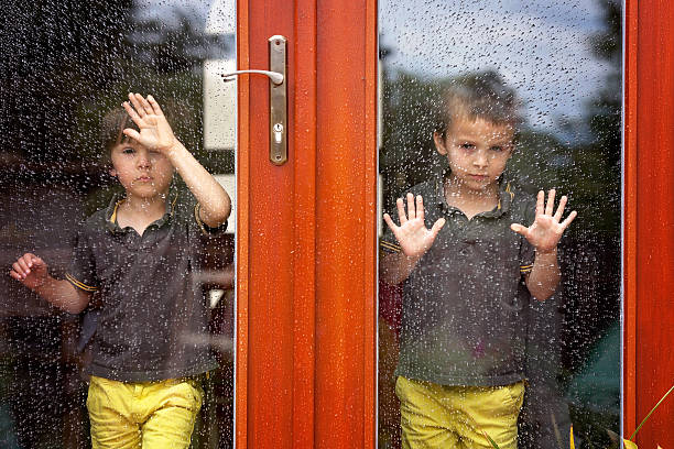 Two little boy, wearing same clothes looking through a big glass door the rain outdoor, summertime