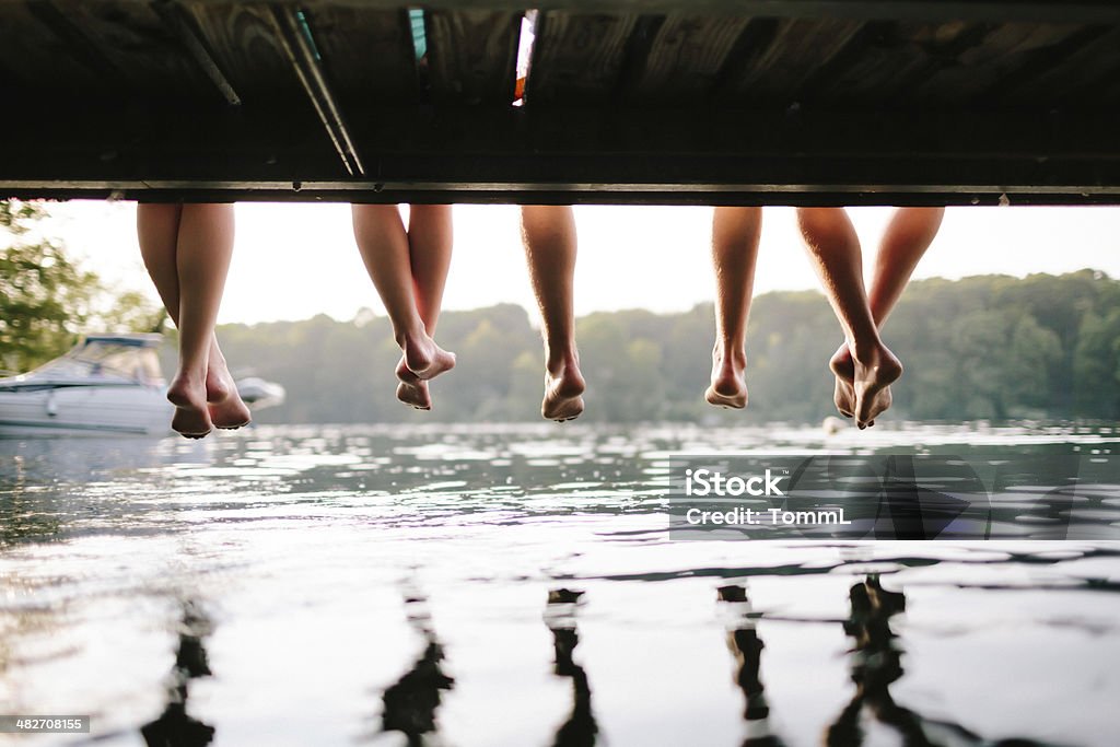 Two Couples Sitting On A Jetty Legs of two couples sitting on a jetty hanging down to the water. Shot from under the jetty at sunset. Summer Stock Photo